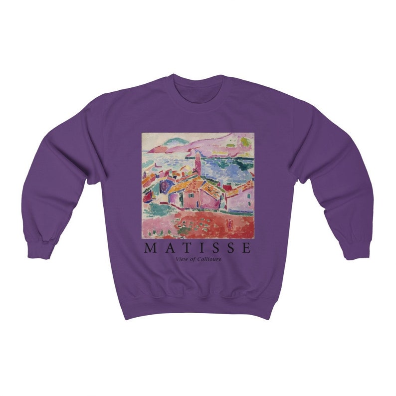 May include: A purple sweatshirt with a printed image of a coastal landscape painting. The artwork shows a town with red-tiled roofs, the sea, and mountains. The text "MATISSE View of Collioure" is printed below the painting in black.
