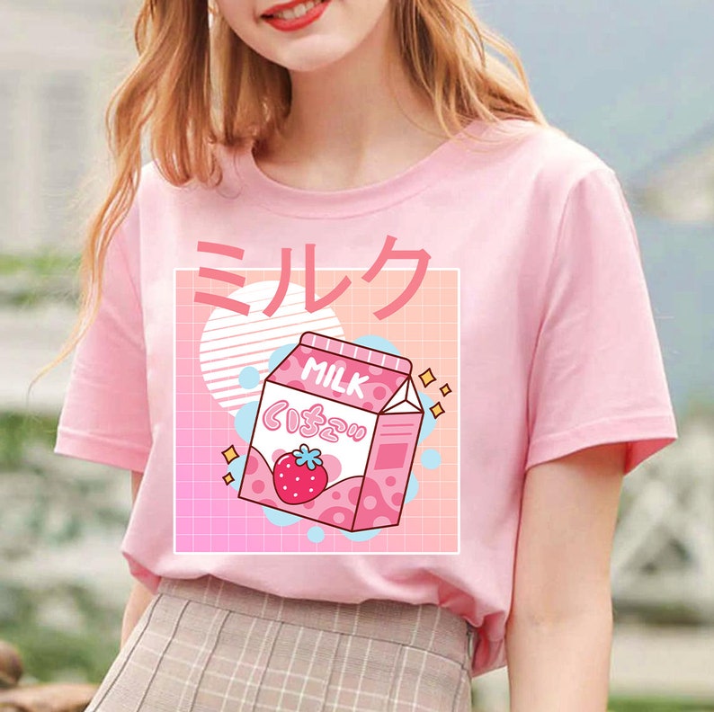 Op de afbeelding: Zachtroze T-shirt met een aardbeienmelkpak-afbeelding en Japanse tekst. Het pak is roze en wit met een aardbeienillustratie. De achtergrond bevat een roze en perzik rasterpatroon met blauwe en witte cirkels.