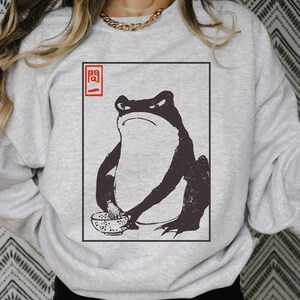 May include: A gray sweatshirt with a black and white illustration of a frog eating ramen noodles. The illustration is framed in a red square with the Japanese character "開" in red.