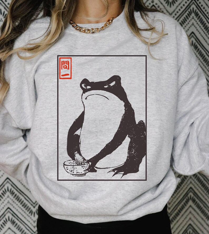 May include: A gray sweatshirt with a black and white illustration of a frog eating ramen noodles. The illustration is framed in a red square with the Japanese character "開" in red.