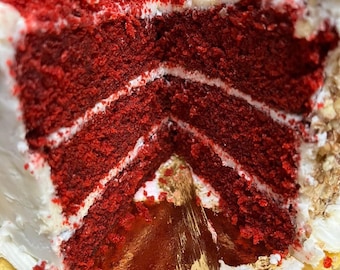 May include: A slice of red velvet cake with white frosting and a crumb topping. The cake has multiple layers with white frosting between each layer. The cake sits on a gold-colored cardboard cake board.