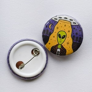 May include: A purple and white button with a pin on the back. The front of the button features a cartoon alien with a green head and a yellow body standing in front of a yellow and orange UFO. The background is a dark blue night sky with stars and a crescent moon.