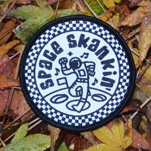 May include: A black and white round patch with a chequered border. The centre of the patch features a cartoon astronaut dancing with the text "Space Skankin'" around the astronaut.