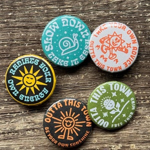 May include: Four round button badges with different designs and sayings. The first badge is green with a white snail and the text "Slow Down Take It Easy". The second badge is white with an orange sun and the text "Take Your Own Advice Outta This Town". The third badge is black with an orange sun and the text "Radiate Your Own Energy". The fourth badge is green with a white flower and the text "Outta This Town Free Your Mind".
