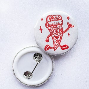 May include: A white button with a red cartoon pizza wearing a Santa hat and the word "RAD" on it. The button has a silver pin on the back.