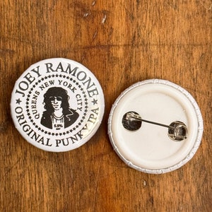 May include: White button with a black and white image of Joey Ramone, the text "Joey Ramone Original Punk IPA", and the words "New York City" in a circle around the image. The back of the button is white with a silver pin.