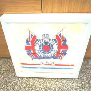 May include: Framed artwork featuring the Rangers Football Club logo, with a red lion in the center, surrounded by a blue and white circular design. Two Union Jack flags flank the logo. The word "DAD" is spelled out at the bottom.
