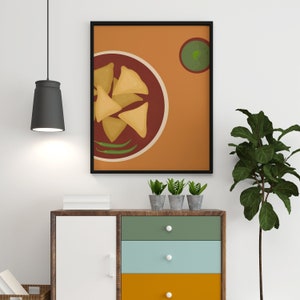 May include: A framed print of a plate of samosas with a side of green chutney. The print is on a brown background with a black frame. The print is hanging on a white wall above a wooden dresser with three drawers.
