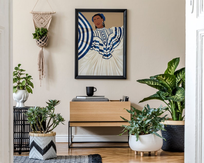 May include: A framed illustration of a woman wearing a blue and white striped dress with a blue headband. The illustration is hanging on a beige wall above a wooden shelf with a black mug, books, and a potted plant.