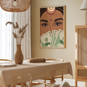May include: A framed illustration of a woman with a bindi and jewelry covering her eyes with US dollar bills. The illustration is set against a beige wall in a dining room with a wooden table, wicker chairs, and a wicker vase with dried flowers.