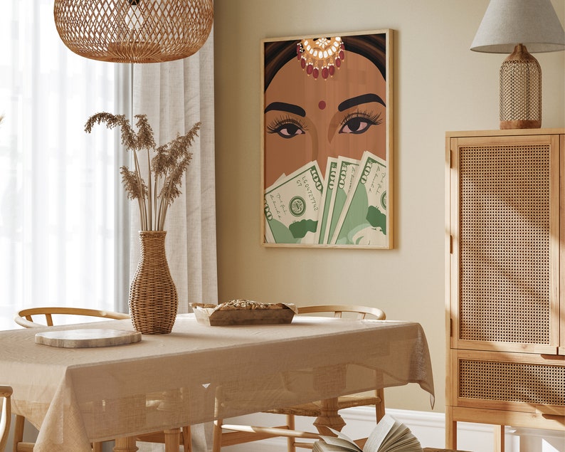 May include: A framed illustration of a woman with a bindi and jewelry covering her eyes with US dollar bills. The illustration is set against a beige wall in a dining room with a wooden table, wicker chairs, and a wicker vase with dried flowers.