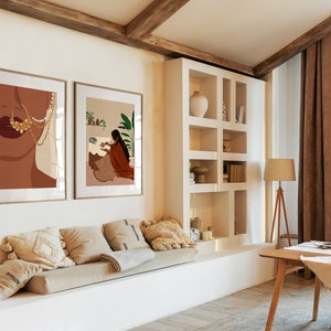 May include: Two framed illustrations of women in a living room with a white couch, a wooden table, and a bookshelf. The illustrations are in a minimalist style with brown, white, and gold colors.