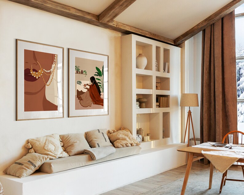 May include: Two framed illustrations of women in a living room with a white couch, a wooden table, and a bookshelf. The illustrations are in a minimalist style with brown, white, and gold colors.