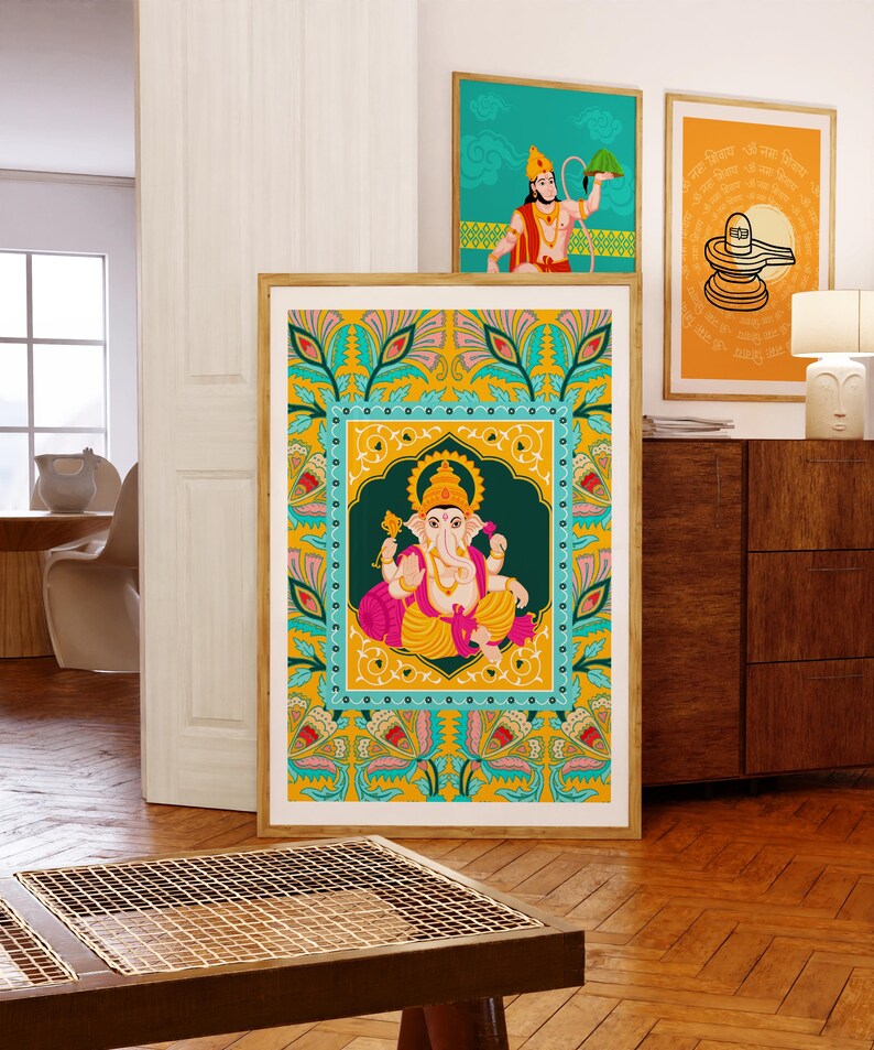 Może przedstawiać: Oprawiony wydruk hinduskiego b&oacute;stwa Ganesha, przedstawiony w żywych kolorach, dominuje na pierwszym planie. Dwa inne oprawione wydruki, z hinduskimi obrazami, są widoczne w tle. Grafika jest wystawiona w dobrze oświetlonym pomieszczeniu z drewnianymi meblami i podłogami.