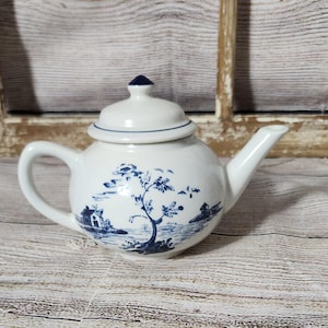 May include: A white teapot with a blue and white floral design. The teapot has a round body and a spout on the side. The teapot is sitting on a wooden surface.