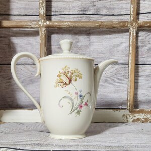 May include: Cream-colored ceramic coffee pot with a lid and a curved handle. The pot features a floral design in shades of yellow, brown, pink, and blue. The pot is set against a rustic wooden window frame.