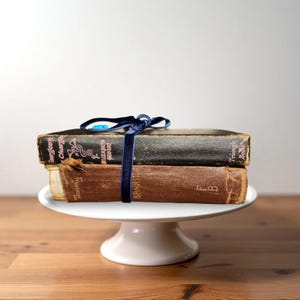 May include: Two antique books, one black and one brown, are stacked on a white cake stand. A dark blue velvet ribbon ties the books together. The books have aged, textured covers and are displayed on a wooden surface.