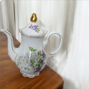 May include: White porcelain teapot with a gold trim and floral design. The teapot has a lid with a gold knob and a handle. The flowers are white, purple, and pink.