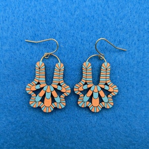 Könnte beinhalten: Ein Paar dekorative Ohrringe mit einem einzigartigen Design. Die Ohrringe haben ein hellorangefarbenes und blaues Muster mit goldfarbenen Haken. Die Ohrringe sind aus Holz gefertigt und haben eine blütenartige Form.