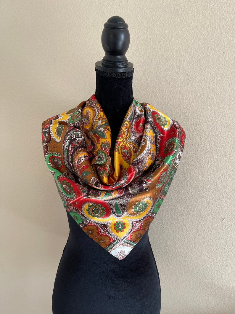 Pu&ograve; includere: Una sciarpa paisley colorata nei toni del marrone, oro, rosso e verde. La sciarpa quadrata &egrave; drappeggiata su un manichino nero, mettendo in risalto il suo motivo intricato e i colori vivaci. Il design della sciarpa presenta un motivo floreale ripetuto.