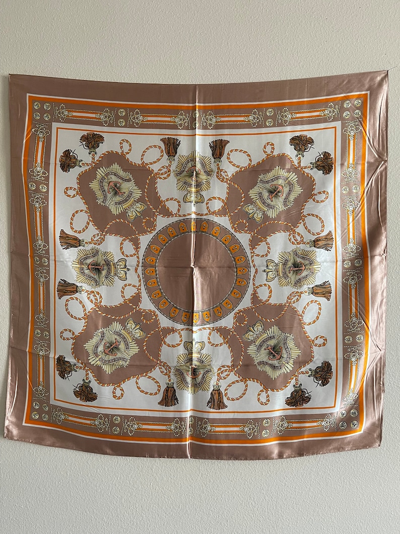May include: A square silk scarf with a brown background and a white center. The center features a repeating pattern of gold rope, tassels, and a stylized design. The scarf has a gold and orange border.