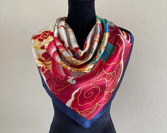 Può includere: Un foulard di seta rosso, bianco, blu e oro con un motivo floreale e nuvole. Il foulard è piegato e drappeggiato attorno a un manichino nero.