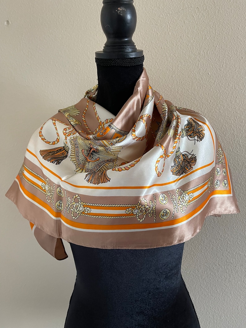 May include: A white silk scarf with a brown and orange pattern of nautical ropes and anchors. The scarf is draped around a black mannequin.