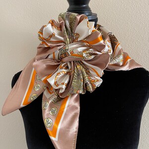 May include: A brown and white silk scarf with a nautical theme. The scarf features a pattern of ropes, anchors, and a gold-colored design. The scarf is tied around a black mannequin's neck.