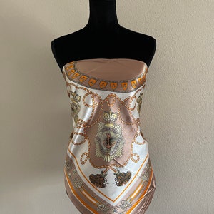 May include: A brown and white silk scarf with a nautical theme. The scarf features a pattern of ropes, anchors, and crowns. The scarf is on a mannequin.
