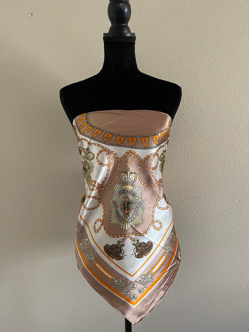 May include: A brown and white silk scarf with a nautical theme. The scarf features a pattern of ropes, anchors, and crowns. The scarf is on a mannequin.