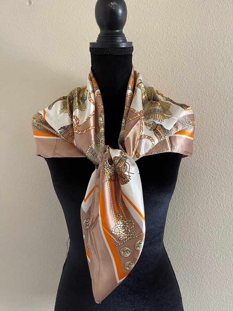 May include: A brown and white silk scarf with a pattern of gold ropes and buttons. The scarf is tied around a black mannequin's neck.