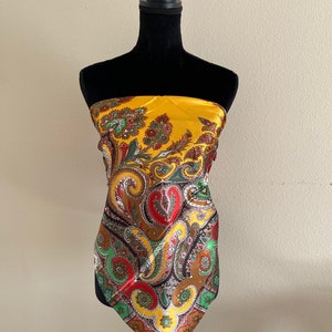 Pu&ograve; includere: Foulard di seta giallo senape con un motivo paisley rosso, verde, marrone e giallo. La sciarpa &egrave; drappeggiata su un manichino nero. La sciarpa ha una consistenza lucida e liscia.