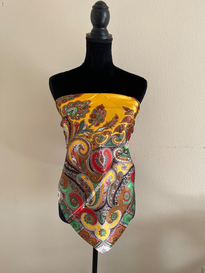 Pu&ograve; includere: Foulard di seta giallo senape con un motivo paisley rosso, verde, marrone e giallo. La sciarpa &egrave; drappeggiata su un manichino nero. La sciarpa ha una consistenza lucida e liscia.
