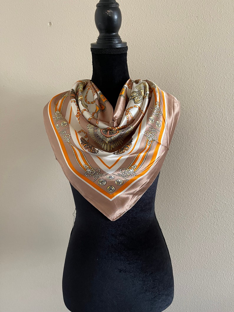 May include: A brown silk scarf with a white and gold pattern of ropes and knots. The scarf is on a black mannequin.