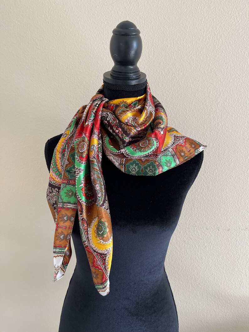 Pu&ograve; includere: Una sciarpa colorata con motivo paisley nei toni del rosso, verde, giallo e marrone &egrave; drappeggiata su un manichino nero. La sciarpa ha una lucentezza setosa e un design dettagliato. La sciarpa quadrata &egrave; piegata e annodata intorno al collo del manichino.