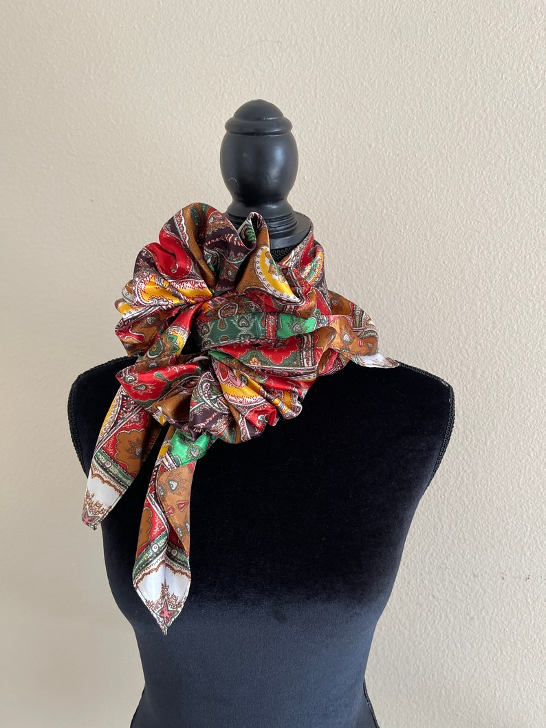 Pu&ograve; includere: Una sciarpa paisley colorata, nei toni del rosso, marrone, giallo, verde e bianco, &egrave; drappeggiata attorno a un manichino nero. La sciarpa &egrave; annodata e disposta per mostrare il suo motivo intricato e i colori vivaci, creando un accessorio decorativo.