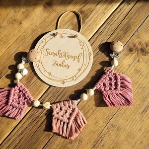 Puede incluir: Un mordedor de madera para bebés con flecos de macramé rosa y blanco y un círculo de madera con el texto "Schlafknopf Zauber" grabado.