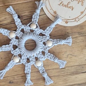 Może przedstawiać: Szary wiszący gwiazdkowy makramowy z drewnianymi koralikami. Gwiazdka jest wykonana z plecionego sznurka i ma okrągły środek. Gwiazdka jest zawieszona na drewnianej powierzchni.