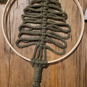 Könnte beinhalten: Ein grüner Makramee-Wandbehang in Form eines Weihnachtsbaums mit einem Quasten am unteren Ende. Der Baum hängt an einem Holzreifen.
