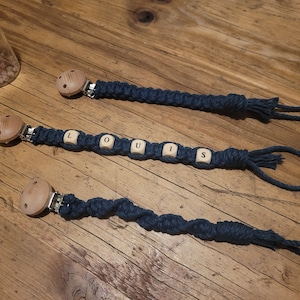 Könnte beinhalten: Drei blaue und weiße geknotete Schnullerketten mit Holzclips und Perlen. Die mittlere Schnullerkette hat den Namen "LOUIS" in Holzperlen geschrieben.