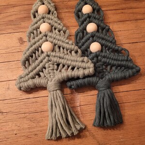 Può includere: Due decorazioni per l'albero di Natale in macramè, una in verde e una in verde scuro, con perline di legno e un anello in cima per appenderle.