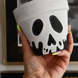Könnte beinhalten: Weißer Keramik-Blumentopf mit einem schwarzen Totenkopf-Design bemalt. Der Totenkopf hat an den Rändern tropfende schwarze Farbe.