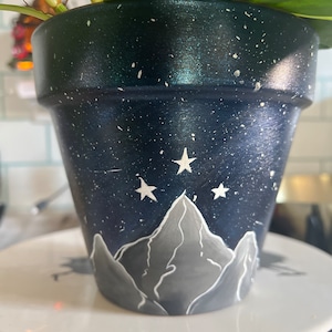 Puede incluir: Una maceta de terracota azul oscuro y verde con un cielo nocturno estrellado blanco y una escena de montaña pintadas sobre ella.