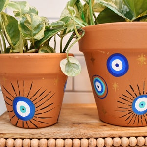 Puede incluir: Dos macetas de terracota con diseños de ojo turco azul y blanco. La maceta más grande tiene un fondo de purpurina dorada y la maceta más pequeña tiene un diseño de sol negro.
