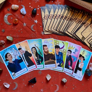 Può includere: Un mazzo di tarocchi con bordi dorati e illustrazioni di persone e simboli. Le carte sono disposte su una superficie rossa con cristalli ed elementi decorativi. Alcune carte sono etichettate con testo come "The World" e "Wands".