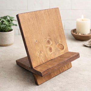 May include: A wooden book stand with a warm, natural finish. The stand is angled for easy viewing and features a small ledge to hold a book or tablet. The wood grain is visible, adding to its rustic charm. A candle and plant are in the background.