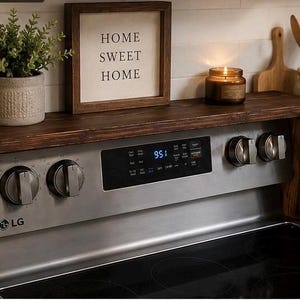 May include: A dark stained wooden shelf installed above a stainless steel LG electric range. The shelf holds a potted plant, a framed sign that reads "HOME SWEET HOME", and a lit candle. Kitchen decor.