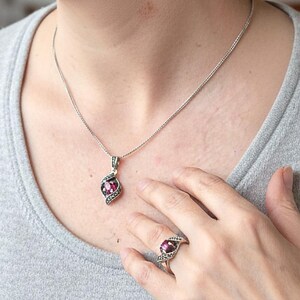 May include: A silver pendant necklace, ring, and earrings set. Each piece features a large, oval, magenta-colored stone surrounded by smaller, decorative stones. The jewelry is worn by a person wearing a gray shirt.