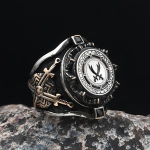 Könnte beinhalten: Ein silberner Ring mit einem schwarzen und weißen kreisförmigen Design mit zwei gekreuzten Schwertern. Der Ring hat eine strukturierte Band und schwarze Edelsteine.
