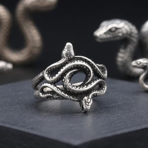May include: A silver-toned metal ring featuring a coiled snake design that wraps around the band. The snake has visible scale textures and two distinct heads meeting at the front, creating an open, intertwined loop shape in the centre.